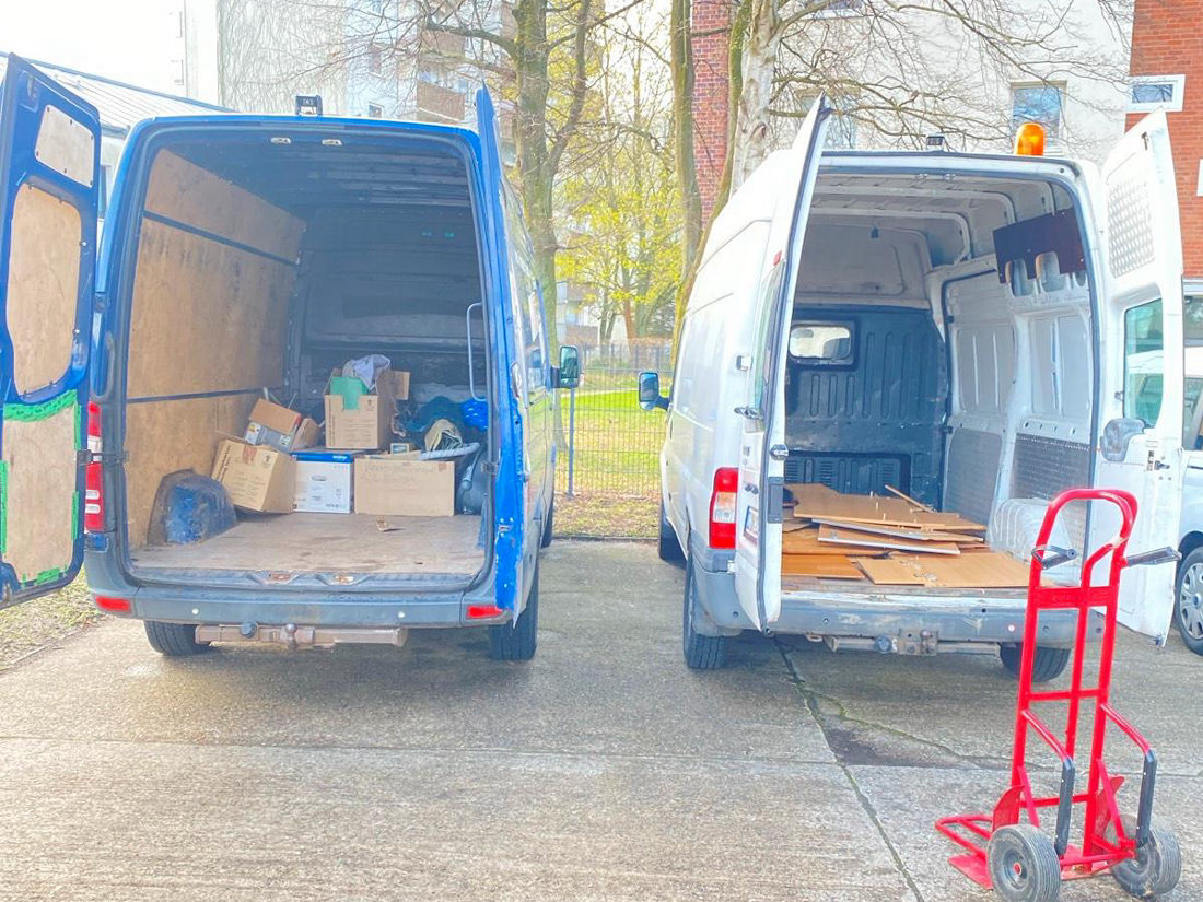Zwei Transporter mit geöffneten Hecktüren auf einem Hof, davor eine Sackkarre